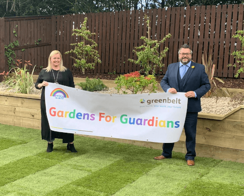 Annie Haslam and Colin Thompson holding up the multicoloured gardens for guardians banner infront of a mini garden containing flora and garden stones